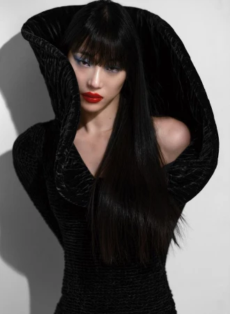 An Asian woman with striking blue eyeshadow and red lipstick wears a textured black hooded gown, shot in a studio with a neutral background.