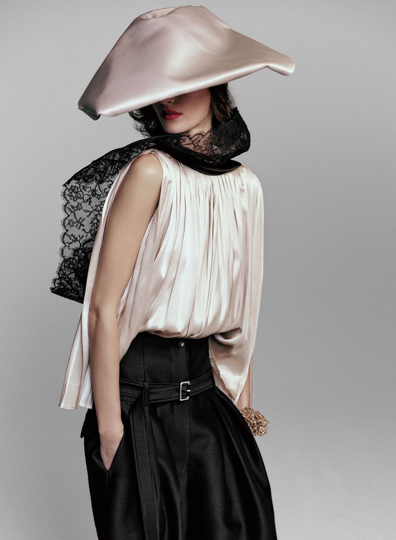Fashion portrait of a woman wearing a silk hat, lace collar, and a satin blouse with a black skirt against a grey studio backdrop.