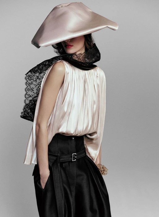 Model wearing a structured silk hat, cream sleeveless pleated top, and black belted trousers against a plain grey backdrop.