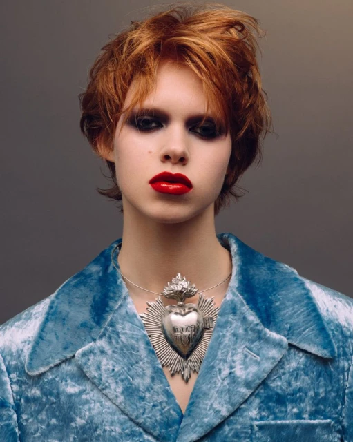 Close-up portrait of a young woman with red hair and bright red lips, wearing a blue velvet jacket and a large silver necklace.