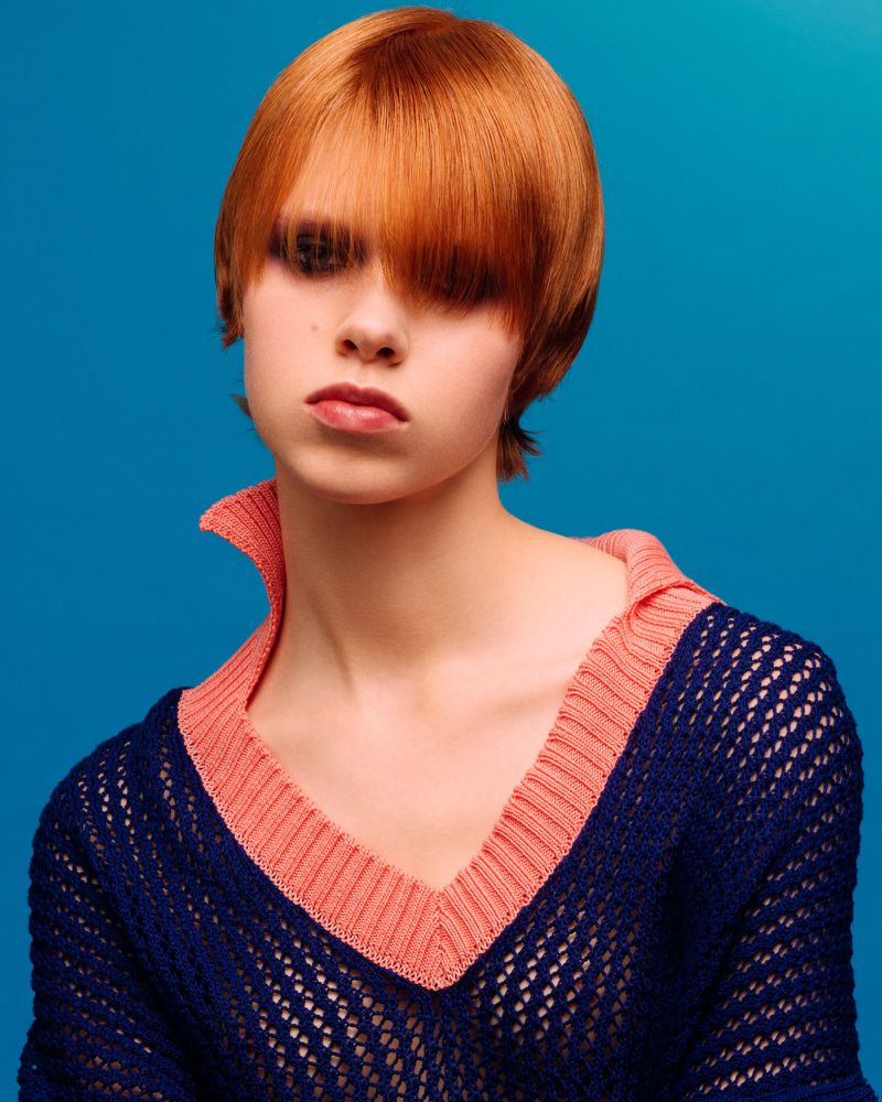 Portrait of a fair-skinned young woman with bright red short hair, wearing a navy and peach-colored net-knit sweater against a blue background.