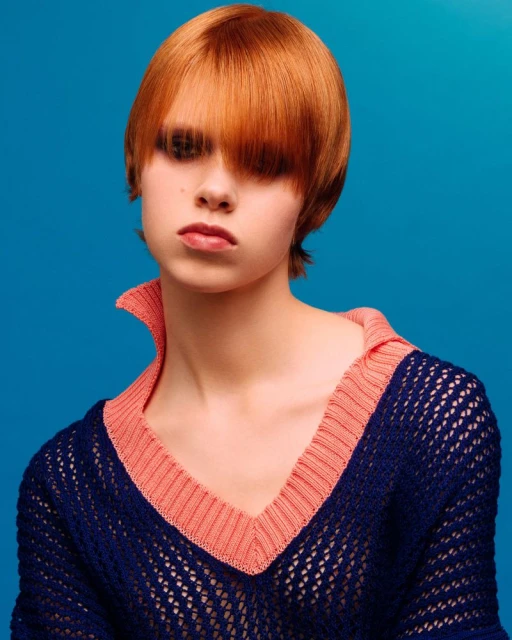 Portrait of a fair-skinned young woman with bright red short hair, wearing a navy and peach-colored net-knit sweater against a blue background.