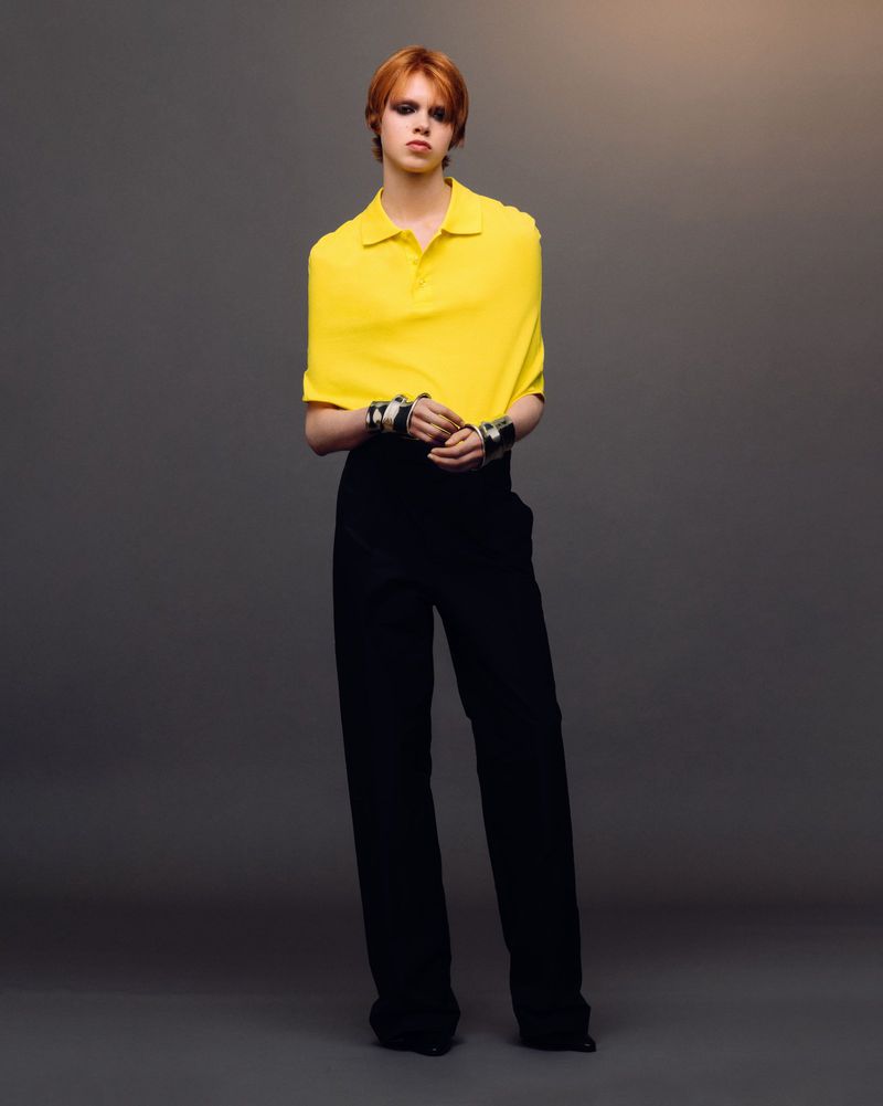 Full-length shot of a red-haired fashion model wearing a yellow polo shirt and black pants against a gradient grey background in studio lighting.