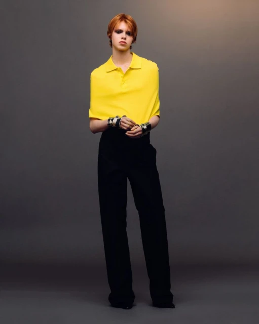 Full-length shot of a red-haired fashion model wearing a yellow polo shirt and black pants against a gradient grey background in studio lighting.