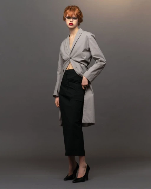 A redhead female fashion model in a blazer and skirt poses in a studio with rim lighting and a neutral gray gradient backdrop.