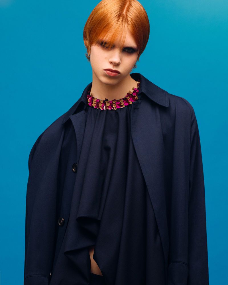 Portrait of a person with red hair wearing a navy trench coat and a frilled collar on a blue backdrop in a studio lighting.