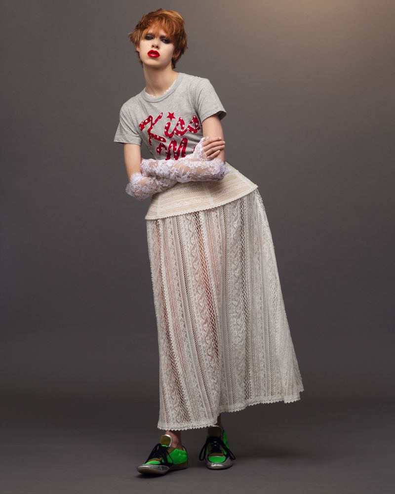A model in a gray t-shirt, white lace skirt, and colorful sneakers poses in a fashion shot against a gray backdrop in a studio.