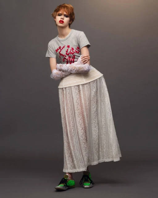 A model in a gray t-shirt, white lace skirt, and colorful sneakers poses in a fashion shot against a gray backdrop in a studio.