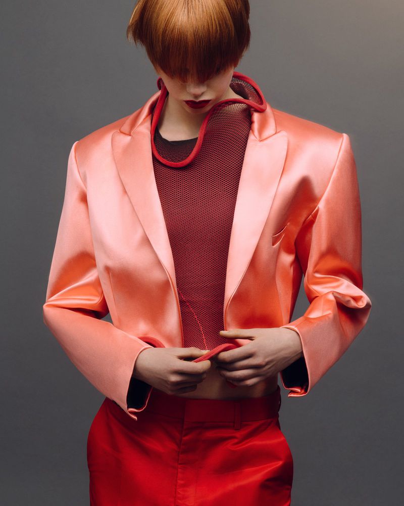 A fashion shot of a model with red hair wearing a peach satin blazer, red top, and pants against a grey backdrop in a studio setting.