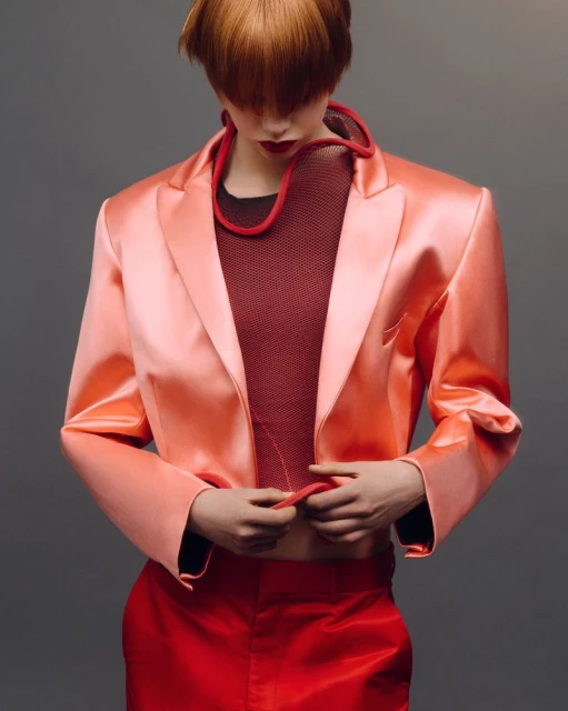 A fashion shot of a model with red hair wearing a peach satin blazer, red top, and pants against a grey backdrop in a studio setting.