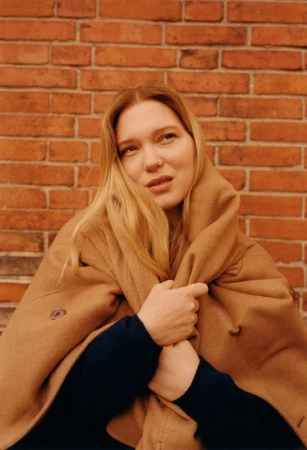 Woman with blonde hair in a tan coat stands in front of a brick wall looking up, creating a portrait style photograph.