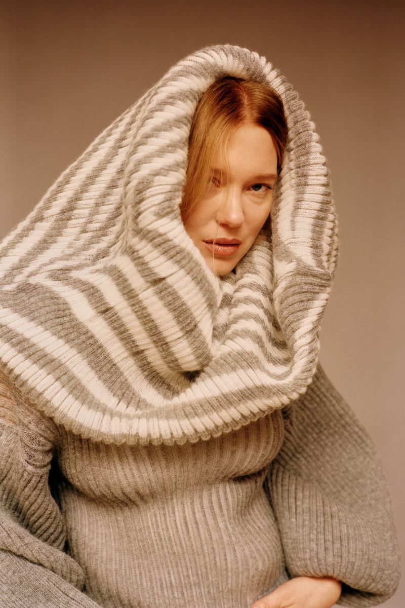 A portrait of a blonde adult female wearing a gray sweater and a large gray and white striped knitted head covering against a neutral background.