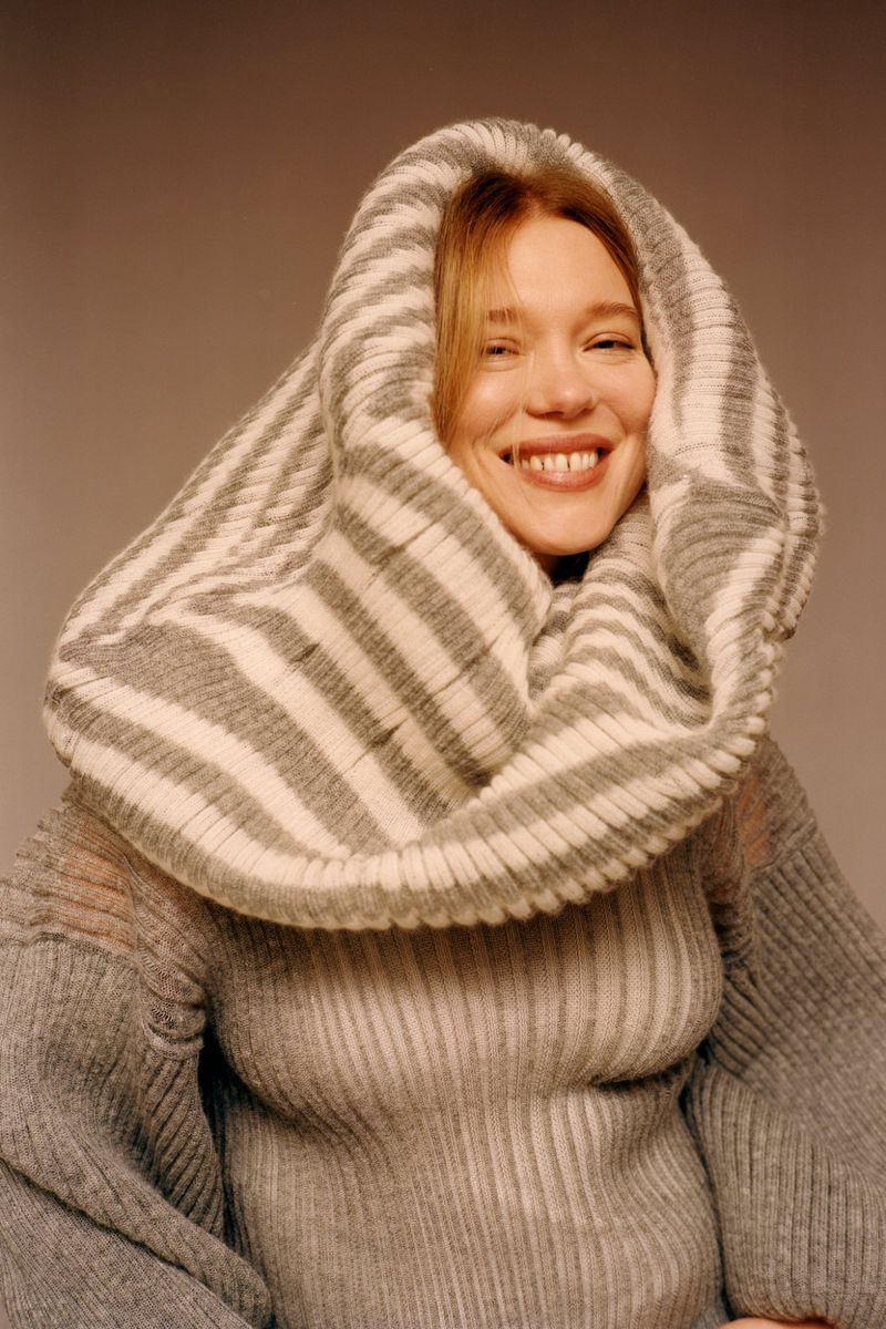 Portrait of a smiling adult woman with fair skin, wearing a ribbed grey sweater and a grey-and-white striped knit hood.