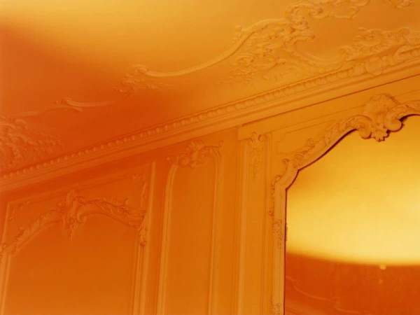 Ornate wall and ceiling with decorative plasterwork, featuring paneling and arches, illuminated by warm, diffused orange and yellow lighting.