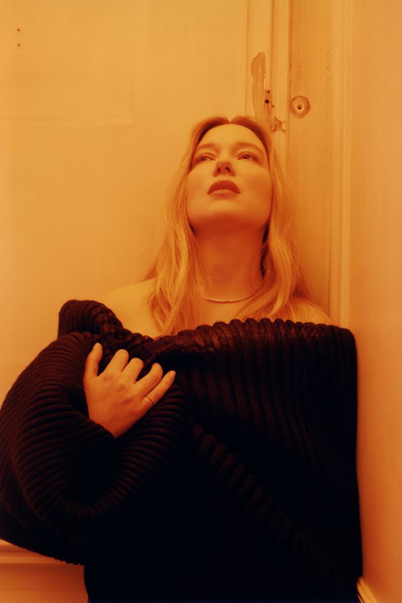 Adult female posing with a black textured garment in an indoor corner setting, orange color grade, looking upwards, with blonde hair.