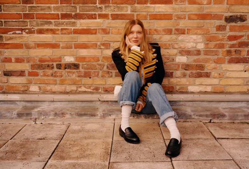 Blonde woman in a black and gold jacket and jeans sits casually against a brick wall on a stone platform wearing black loafers.