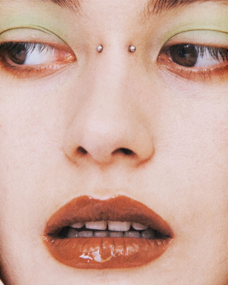 Close-up shot of a young adult female with brown eyes, glossy lips, fair skin and eyebrow piercing looking to the side in studio lighting.