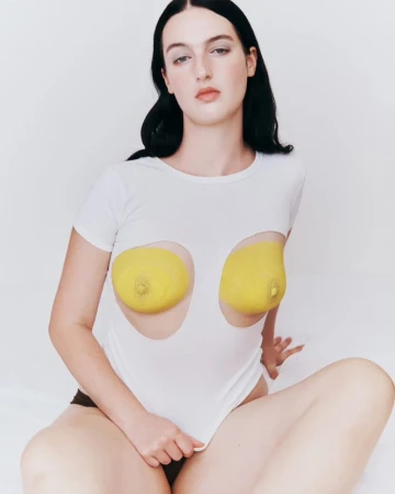Brunette woman in a white bodysuit with yellow breast coverings, seated indoors against a white backdrop, looking at the camera.