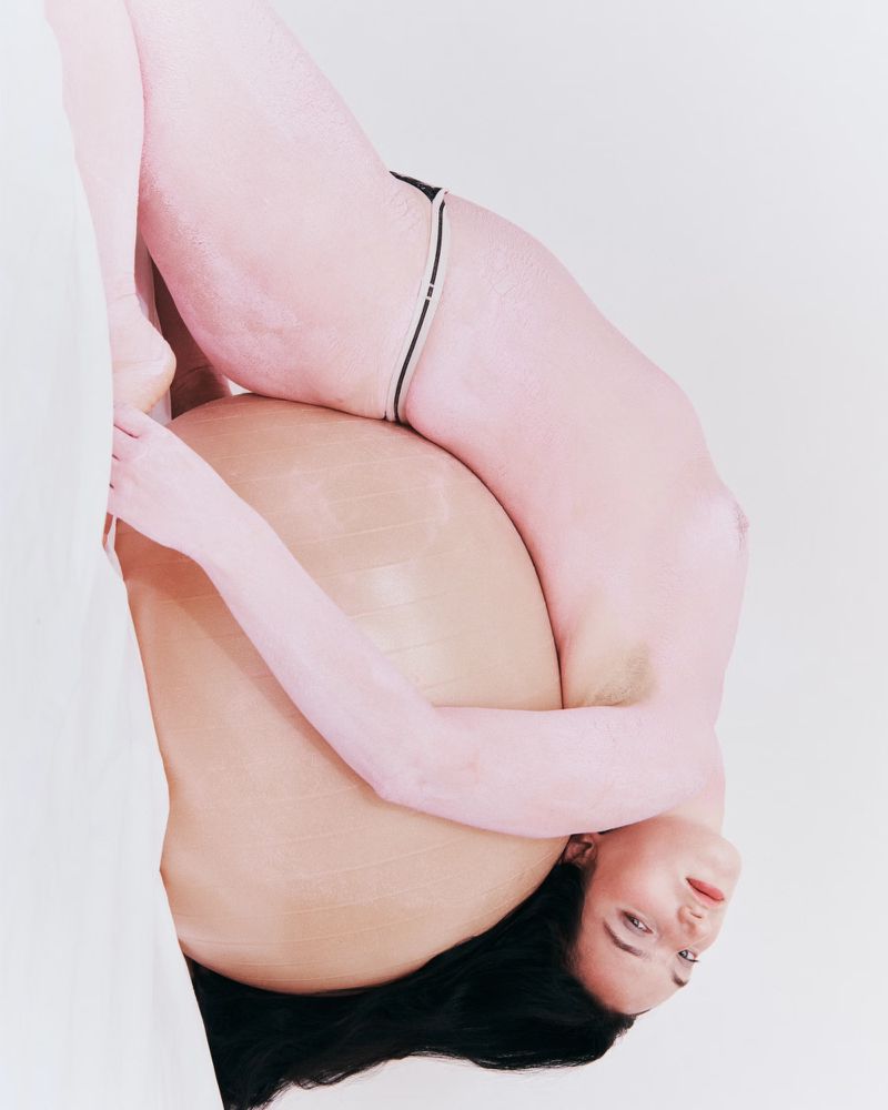 Artistic portrait of a nude woman covered in light pink paint, holding a brown, smooth, round shape. Studio shot on white.