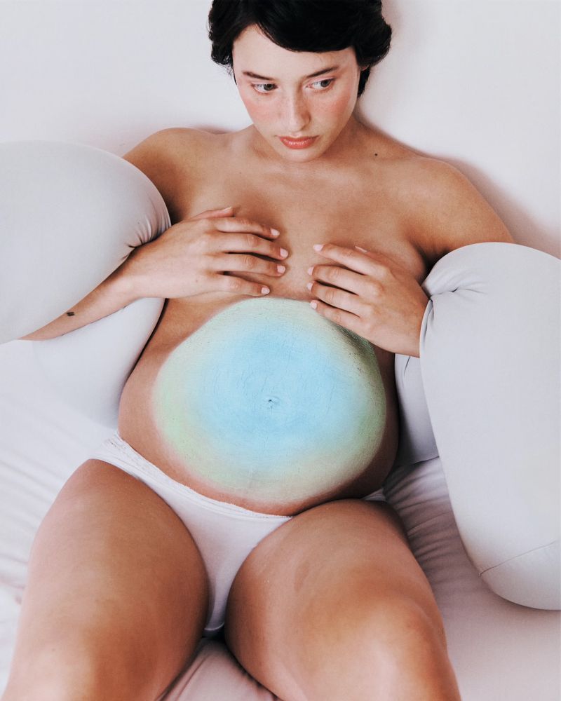 A pregnant woman with a blue and green painted belly sits on a white bed with large, soft, white pillow-like objects.