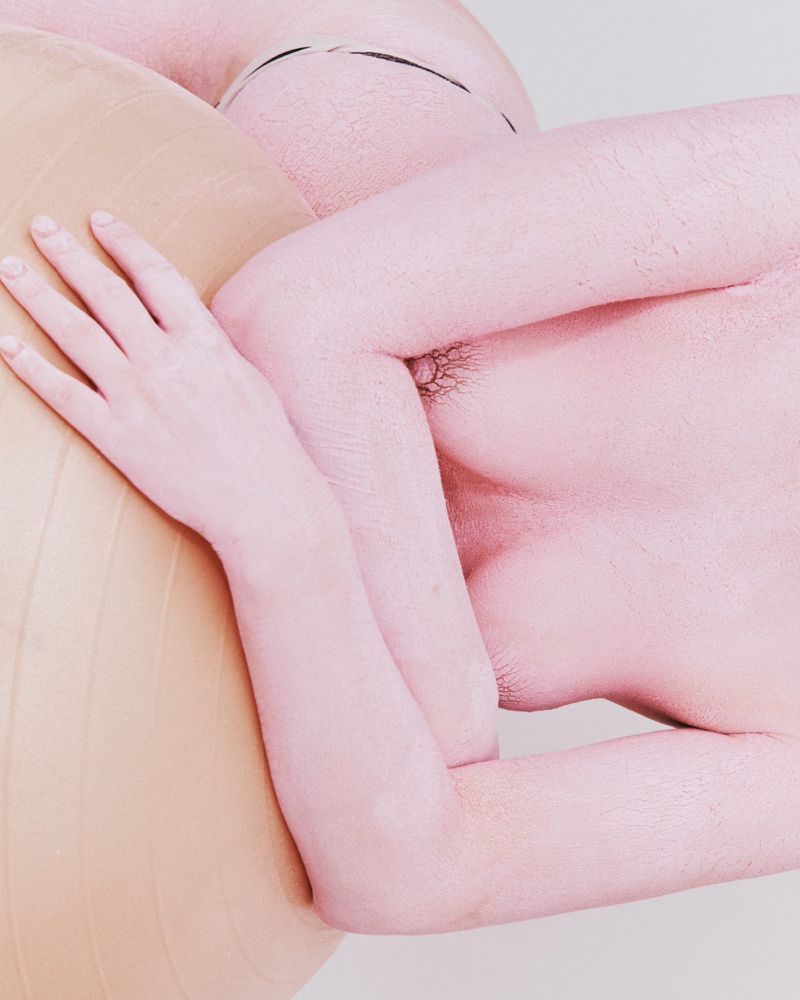 Nude human figure covered in pink paint poses with a beige exercise ball indoors, utilizing soft lighting and a low-angle perspective.