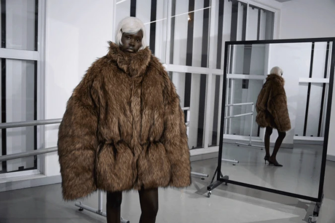 A fashion model wearing a large brown fur coat and white wig poses in a photo studio with a reflective standing mirror.