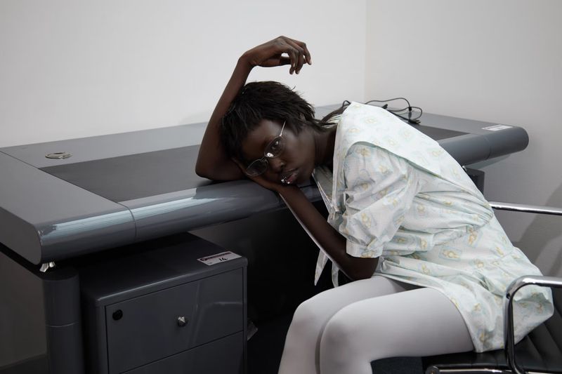 Woman with dark skin and short hair wearing glasses and patterned medical gown rests head on modern grey desk in indoor setting.