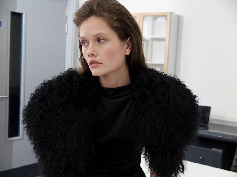Woman in black dress and furry bolero poses indoors with white walls, wearing minimal makeup and with hair neatly styled.
