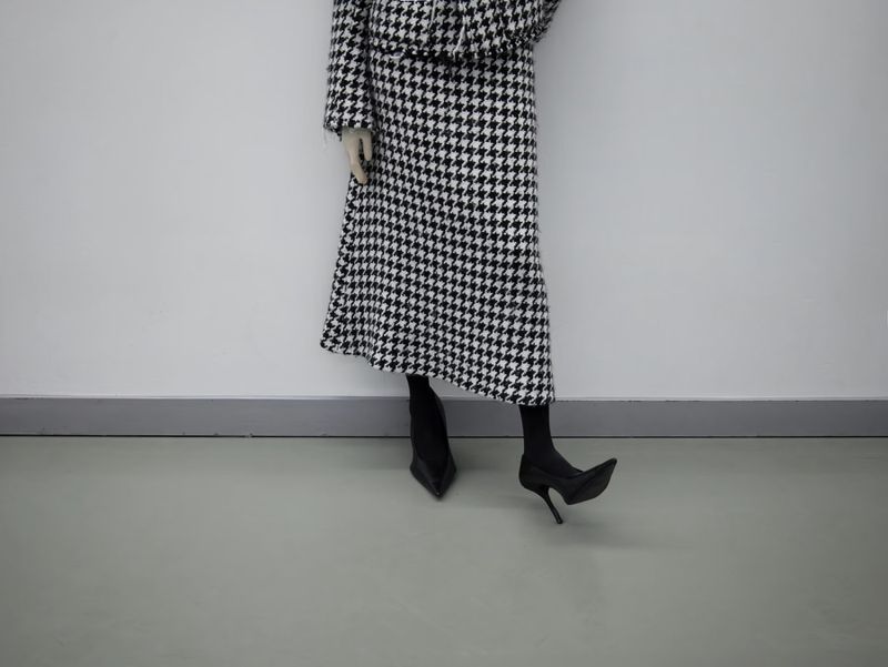 Fashionable woman in a black and white houndstooth jacket and skirt paired with black heels against a white wall backdrop.