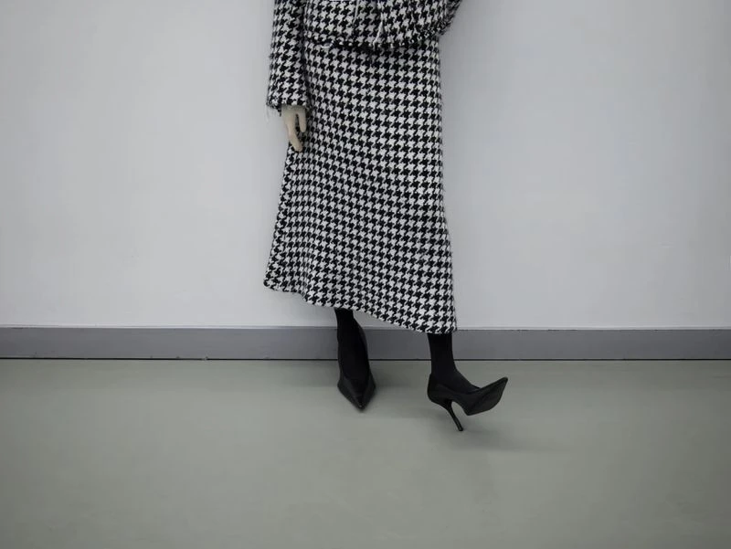Fashionable woman in a black and white houndstooth jacket and skirt paired with black heels against a white wall backdrop.
