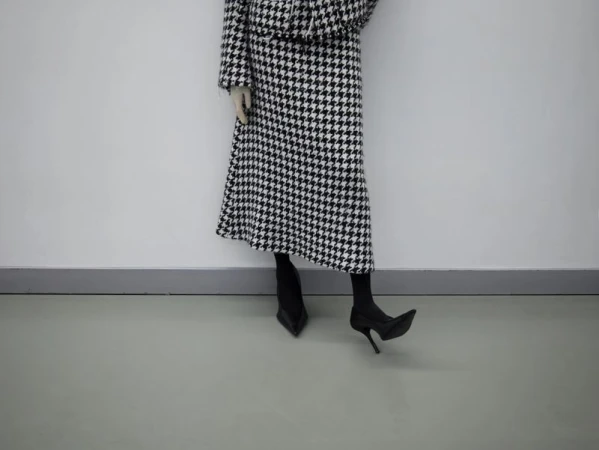 Fashionable woman in a black and white houndstooth jacket and skirt paired with black heels against a white wall backdrop.