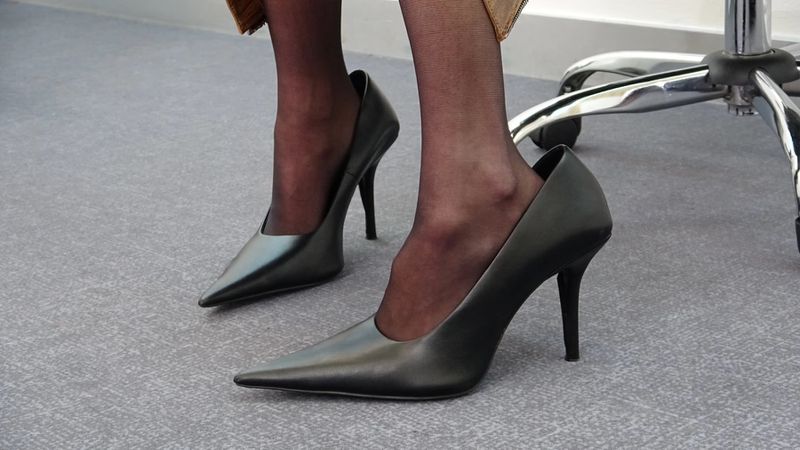 Low-angle shot of black high-heeled shoes on a woman indoors, complemented by sheer black stockings and a stylish gold skirt.