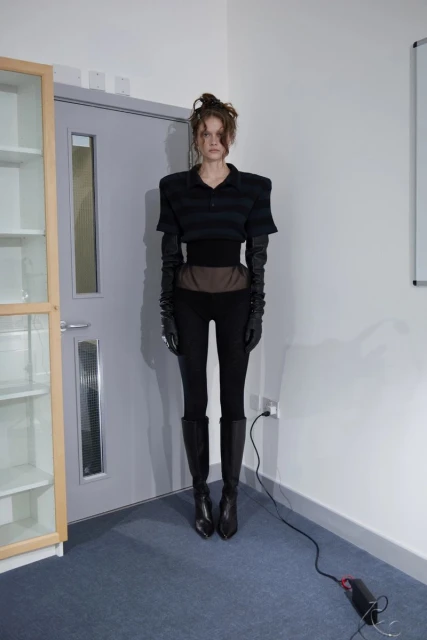 Adult woman in a black polo shirt, black pants, and knee-high boots standing in a room with a cabinet and door in a full shot.