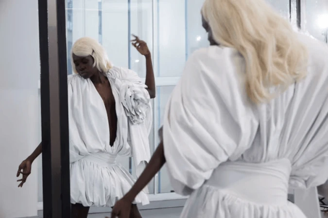 Black model with platinum blonde hair wearing a sculptural white dress posing in front of a mirror