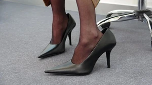 Close-up of a person wearing black pointed-toe high heels and sheer black stockings against a grey office carpet background.