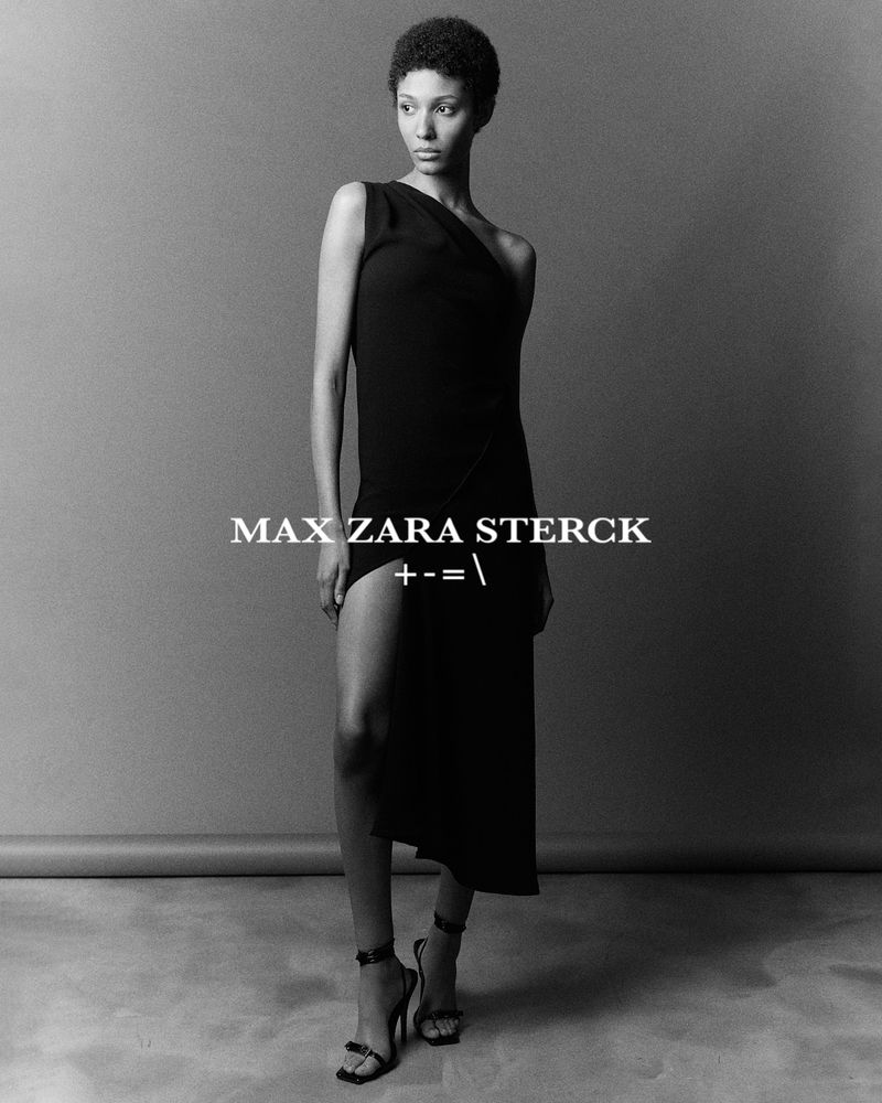 Portrait of a black female model wearing a black, one-shoulder dress and heeled sandals in a studio setting with a gray background.