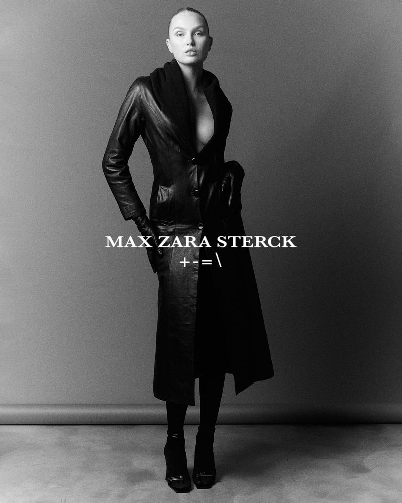 Elegant studio shot of a slender fashion model in a black leather coat, dark stockings, and high heels against a gray backdrop.