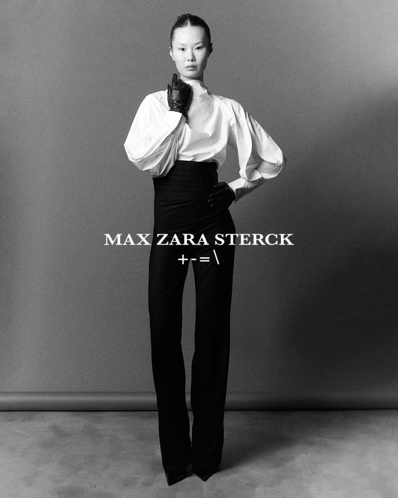 Model in white blouse and black trousers wearing dark gloves against a grey seamless studio backdrop.