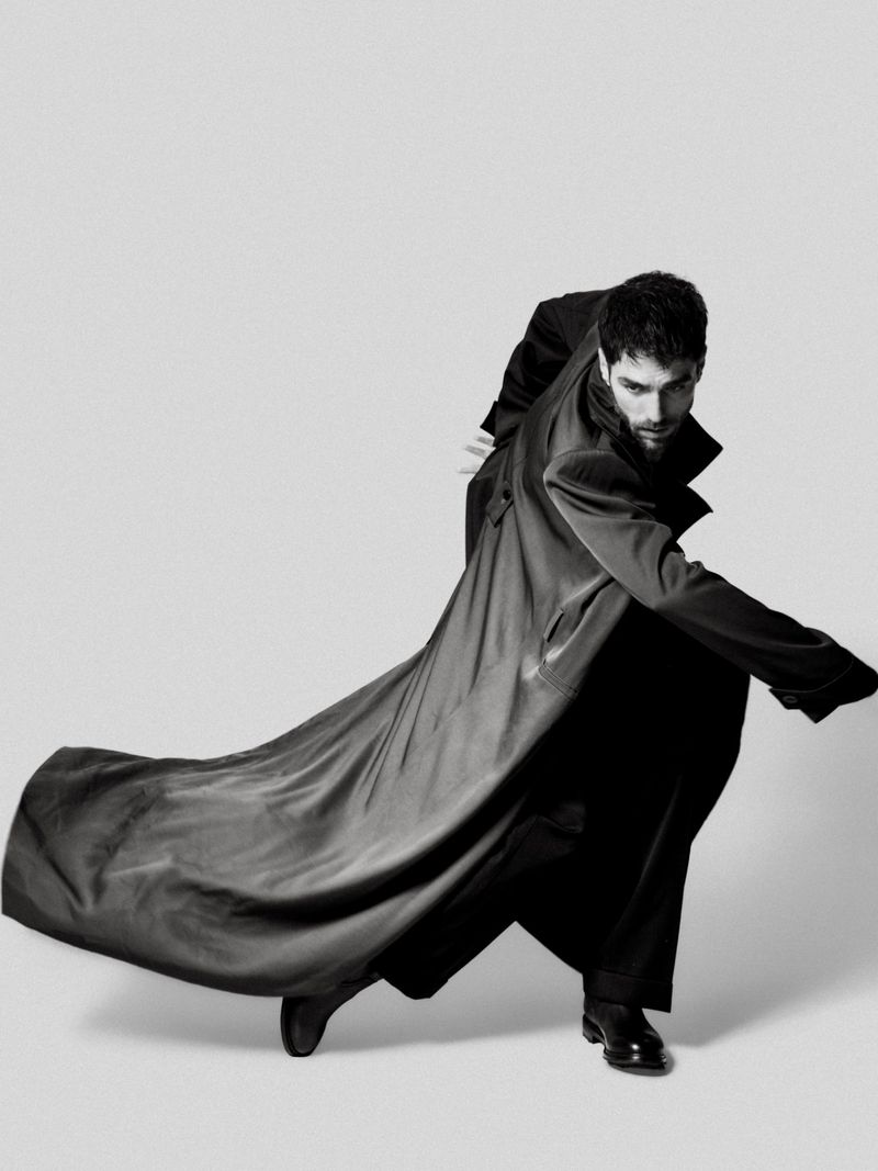 A dark-haired man strikes a dynamic pose in a flowing black coat against a clean white backdrop, showcasing fashion and movement.