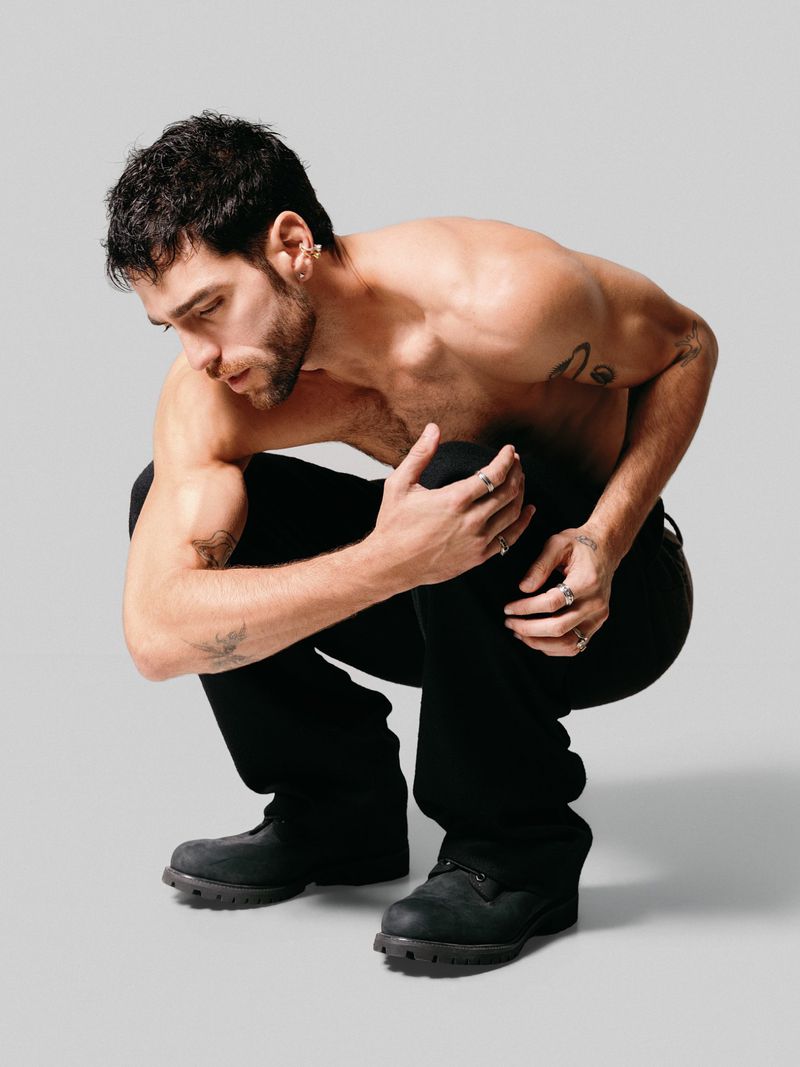 Man in black jeans and work boots squats shirtless against a light gray backdrop, with tattoos and multiple rings on his fingers.