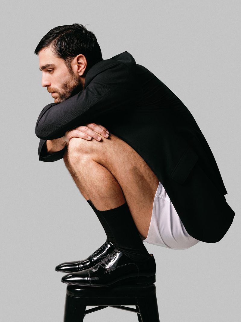 Man squats on a black stool wearing a black blazer, black socks and shiny black shoes, but only white underwear.
