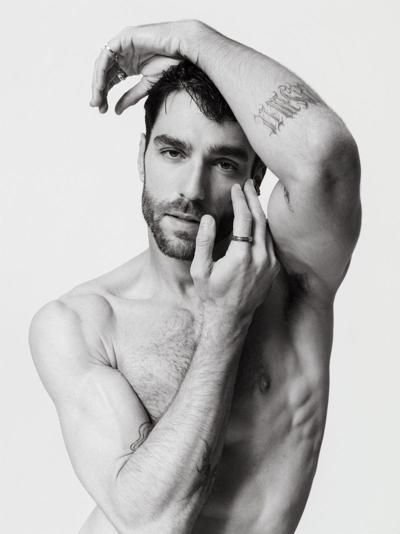 Black and white portrait of a bare-chested man with hands near his face, featuring tattoos and jewelry, set against a plain background.