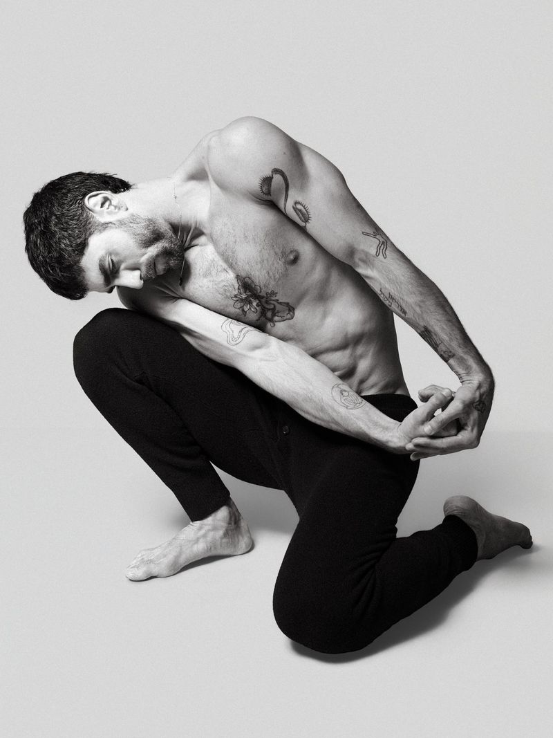 An adult male with tattoos is posing on his knees in a studio. He is wearing black pants and is shirtless in a black-and-white photo.