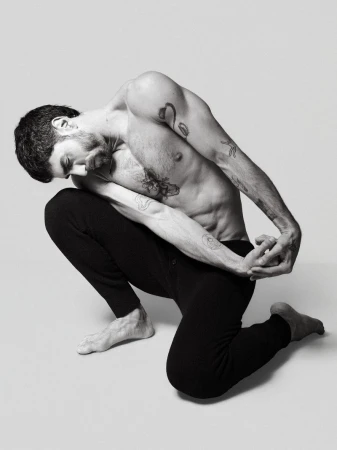 An adult male with tattoos is posing on his knees in a studio. He is wearing black pants and is shirtless in a black-and-white photo.