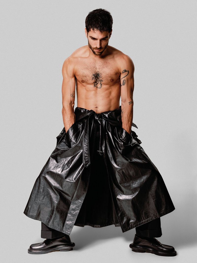 An adult man with a beard stands shirtless, hands in pockets of a long, black, shiny coat, over black pants and platform shoes, against a grey background.