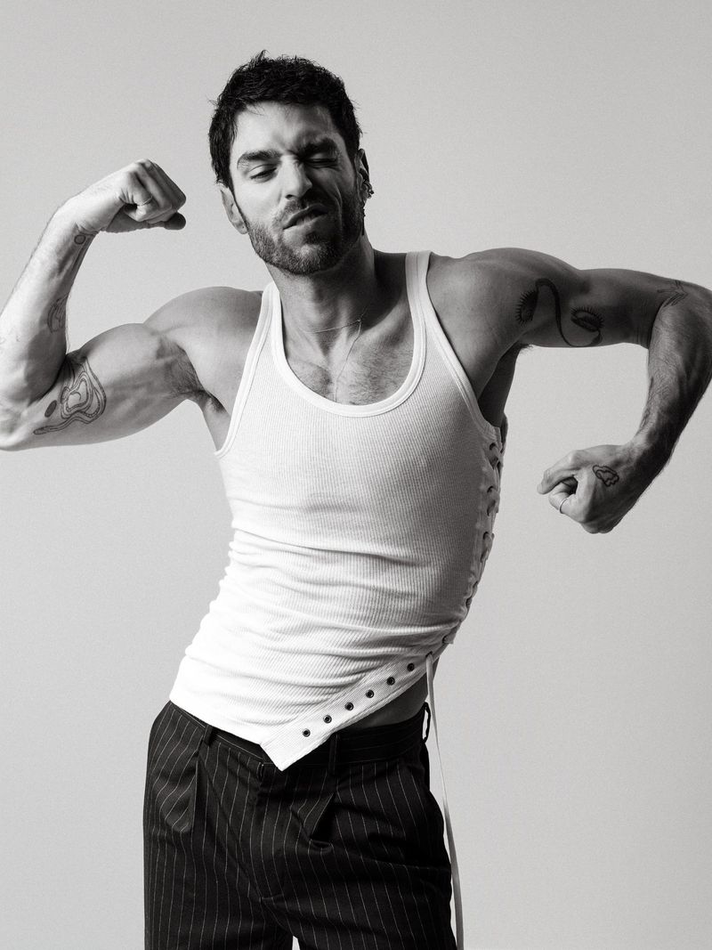 Black and white portrait of a muscular man in a tank top and striped pants flexing his biceps against a plain background in a studio.