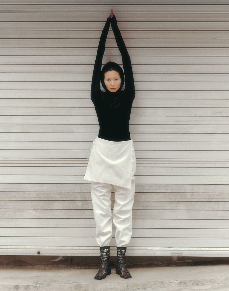 Full-length shot of an adult female raising her arms overhead, wearing a black top, white skirt, white pants, and brown boots, in front of a metal garage door.