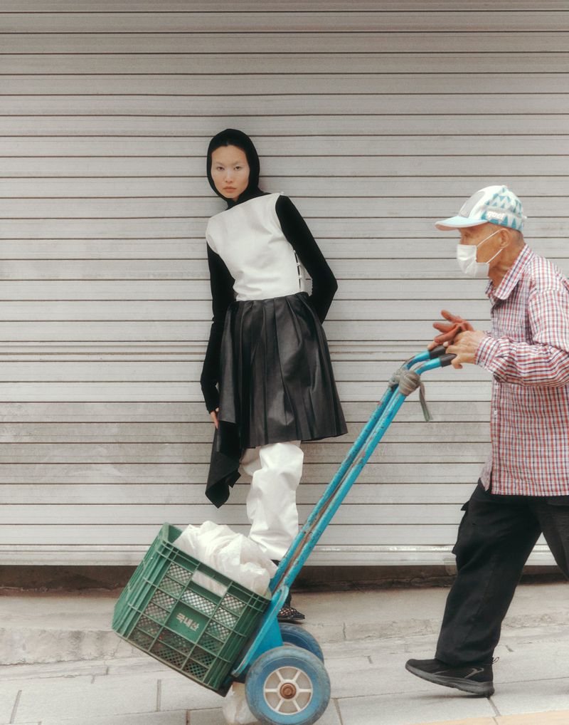 Fashion shot of a young Asian woman in black and white outfit standing next to an older Asian man pulling a cart on a city street.