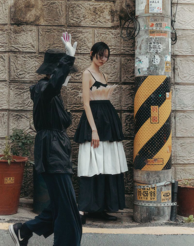 A street-style fashion shot with a model in a tiered dress and a person in a masked costume standing against a textured wall.