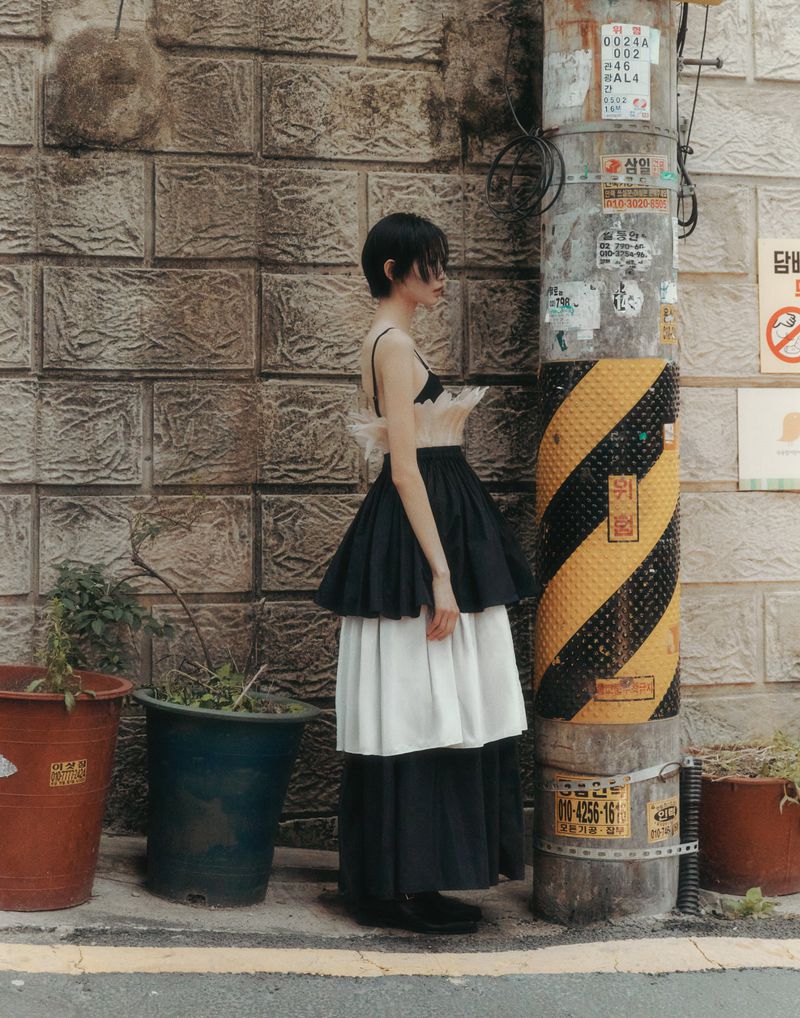 An adult female with short dark hair stands outdoors on a city sidewalk next to a utility pole and a brick wall wearing a layered dress.